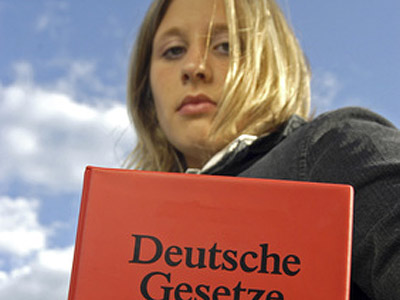 Eine Person hält ein rotes Buch mit dem Titel "Deutsche Gesetze" vor einem wolkigen Himmel.